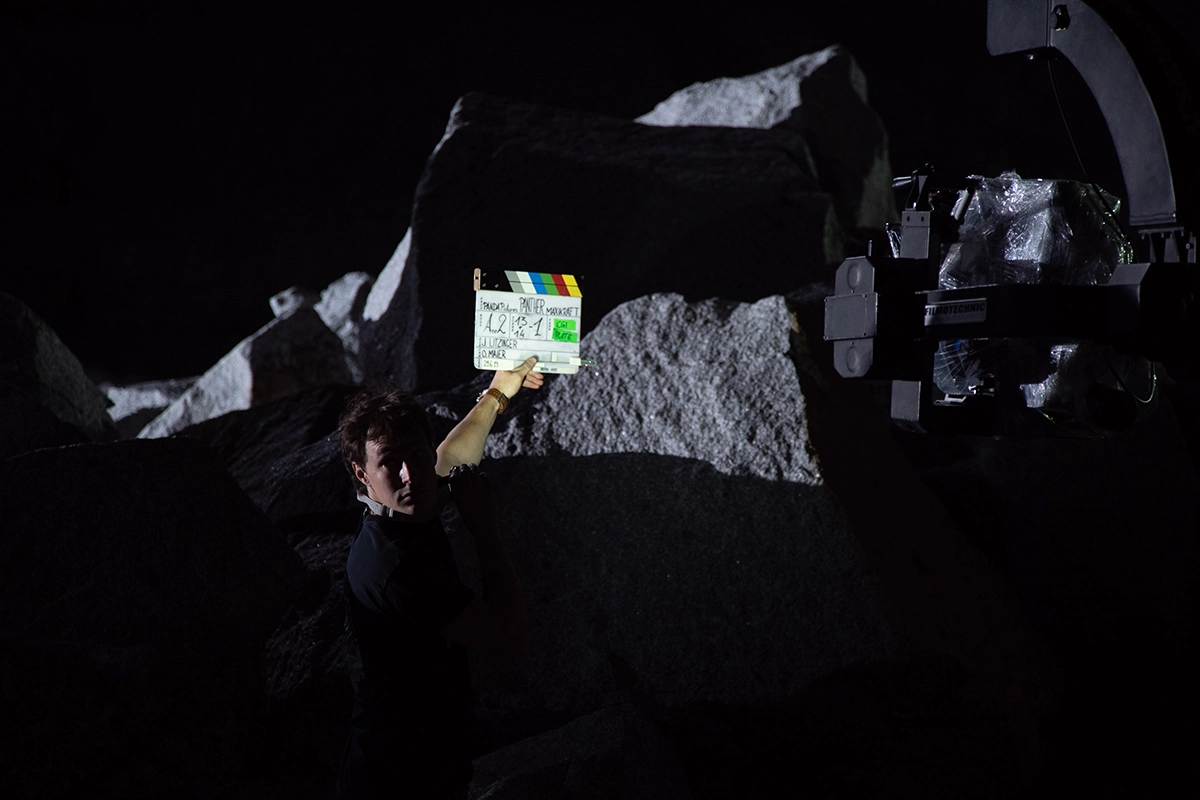 Imagefilm der Filmproduktion Panda Pictures aus München und Ulm für Maxikraft. Ein junger Kameramann oder Assistent hält eine Filmklappe hoch, um die Szene zu markieren, während im Hintergrund Felsen und Filmtechnik zu sehen sind.