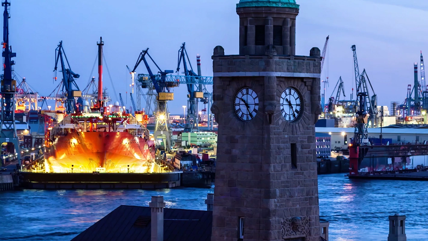 Imagefilm der Filmproduktion Panda Pictures aus München und Ulm für SupplyOn. Ein Uhrturm im Vordergrund des beleuchteten Hamburger Hafens bei Dämmerung, stimmungsvoll eingefangen für eine atmosphärische Aufnahme.