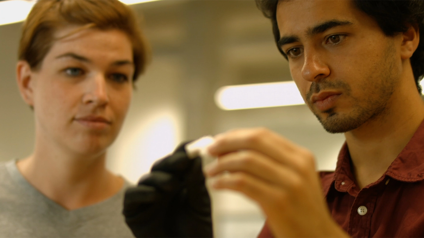 Imagefilm der Filmproduktion Panda Pictures aus München und Ulm für Techfounders. Eine Nahaufnahme zeigt zwei Forscher, einen männlichen und einen weiblichen, die konzentriert ein kleines, weißes Objekt mit schwarzen Handschuhen untersuchen.