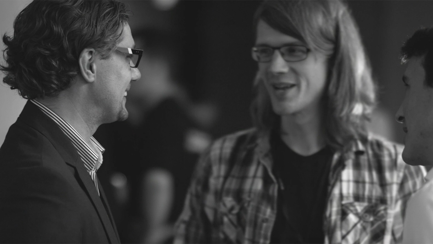 Imagefilm der Filmproduktion Panda Pictures aus München und Ulm für TechFounders. Eine schwarz-weiß gefilmte Einstellung zeigt zwei lachende Darsteller im Gespräch, wobei der Fokus auf dem seitlich stehenden Mann mit Brille und Anzug liegt.