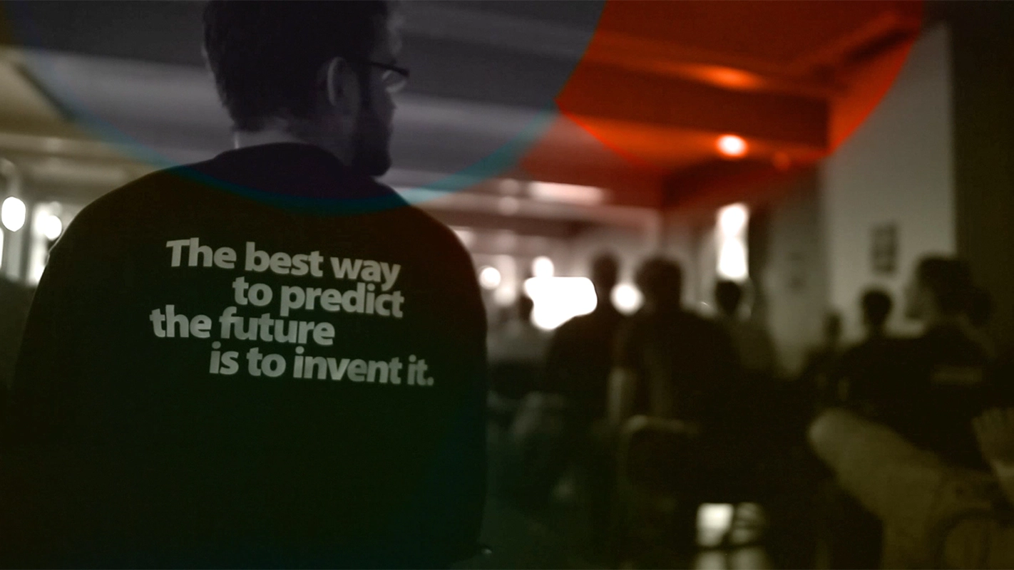 Imagefilm der Filmproduktion Panda Pictures aus München und Ulm für TechFounders. Eine dokumentarische Einstellung von hinten zeigt einen Darsteller mit der Aufschrift „The best way to predict the future is to invent it“ vor verschwommenen Zuhörern.