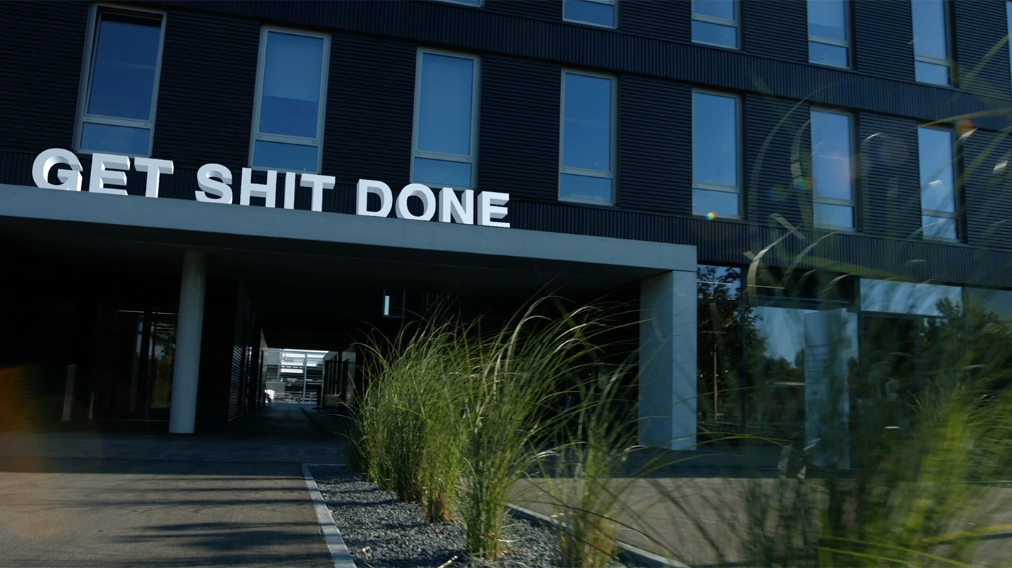 Imagefilm der Filmproduktion Panda Pictures aus München und Ulm für TechFounders. Eine weite Einstellung zeigt das moderne Gebäude von TechFounders mit der klaren Botschaft "GET SHIT DONE" als großen weißen Schriftzug davor, umgeben von grünen Pflanzen.