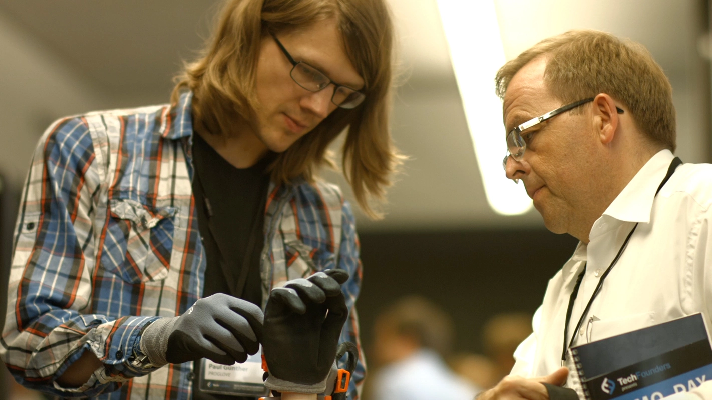 Imagefilm der Filmproduktion Panda Pictures aus München und Ulm für TechFounders. Eine szenische Aufnahme zeigt zwei Darsteller, die sich intensiv mit einem High-Tech-Handschuh beschäftigen; der Fokus liegt auf der Interaktion und dem Gerät.
