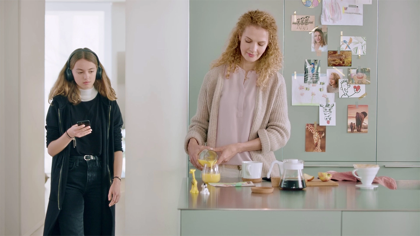 Imagefilm der Filmproduktion Panda Pictures aus München und Ulm für GIRA. Eine Darstellerin mit Kopfhörern und Handy blickt nachdenklich in einem hellen Raum, während eine zweite Darstellerin am Küchentisch steht und Saft einschenkt.