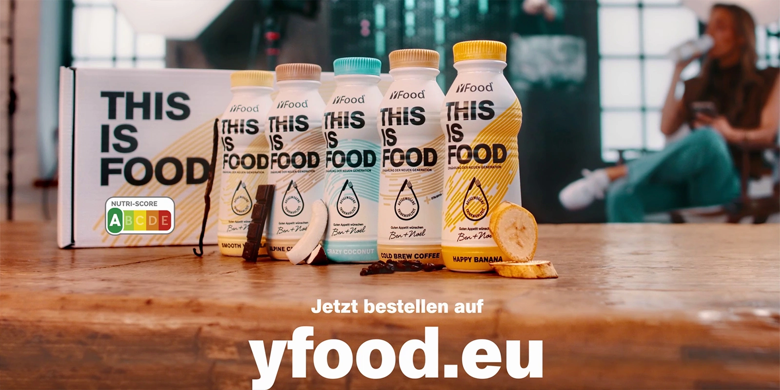 TV Werbefilm der Filmproduktion Panda Pictures aus München und Ulm für yFood. Eine Reihe von yFood-Flaschen in verschiedenen Geschmacksrichtungen mit Nutri-Score auf einem Holztisch, im Hintergrund eine unscharfe Person.