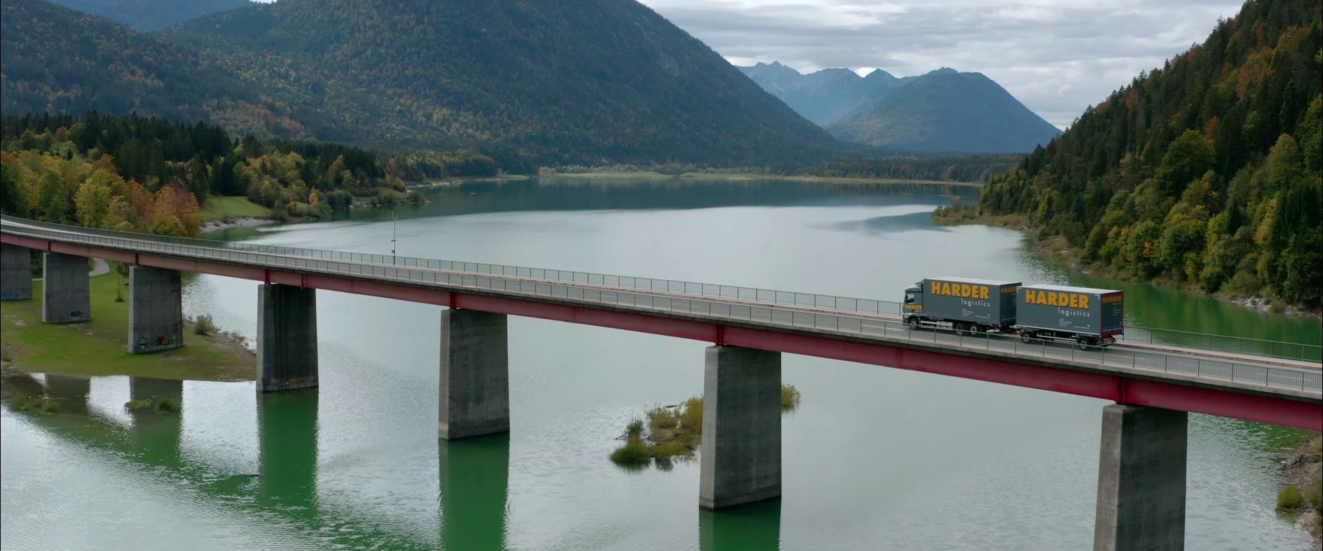 Imagefilm der Filmproduktion Panda Pictures aus München und Ulm für HARDER. Ein Harder-Lkw überquert eine lange Brücke über einen See mit Bergen. Eine beeindruckende Luftaufnahme der logistischen Meisterleistung.