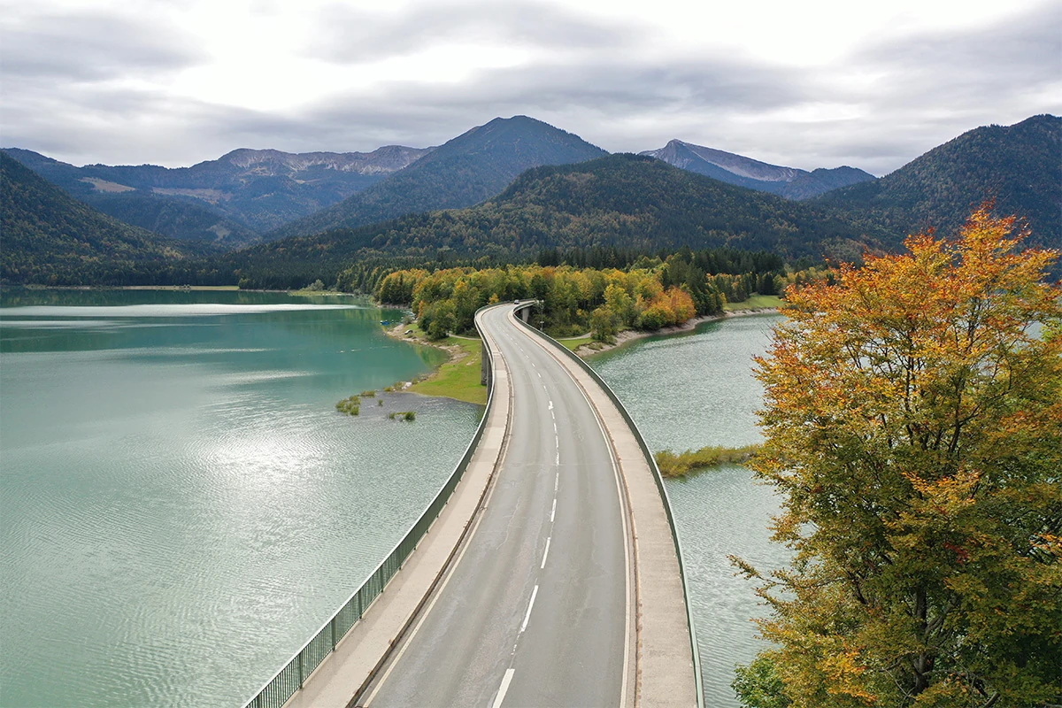 Imagefilm der Filmproduktion Panda Pictures aus München und Ulm für HARDER. Eine gewundene Brücke über einen See, gesäumt von herbstlichen Bäumen und Bergen. Die Bildkomposition erzeugt visuelle Harmonie.