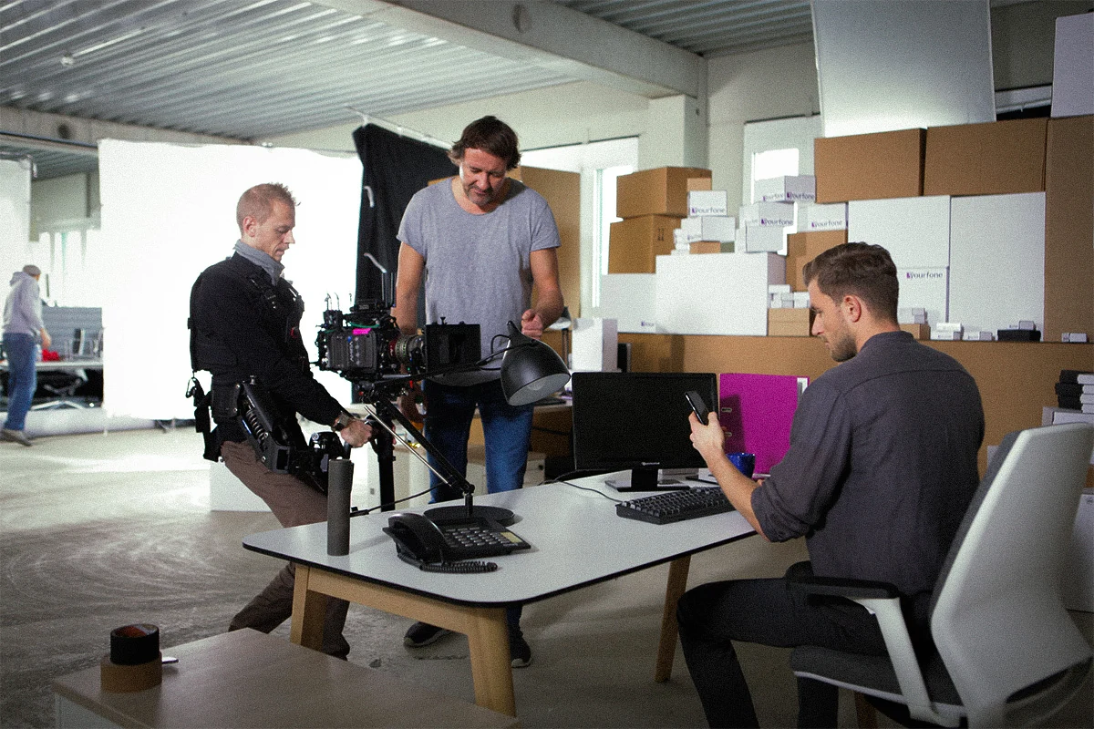 TV Werbespot der Filmproduktion Panda Pictures aus München und Ulm für yourfone. Ein Darsteller sitzt am Schreibtisch, während der Kameramann mit seinem Equipment und ein weiteres Crew-Mitglied die Einstellungen überprüfen.