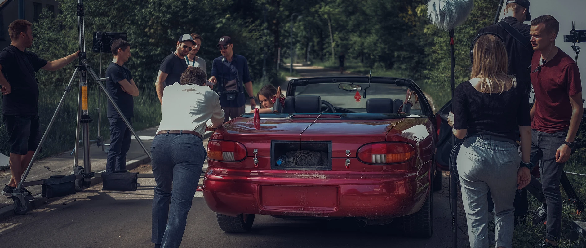 Imagefilm der Filmproduktion Panda Pictures aus München und Ulm für Scheuermann & Heilig. Eine Filmcrew umringt ein rotes Cabriolet an einem Drehort im Freien, während ein Kameramann eine Einstellung mit dem Darsteller im Auto bespricht.
