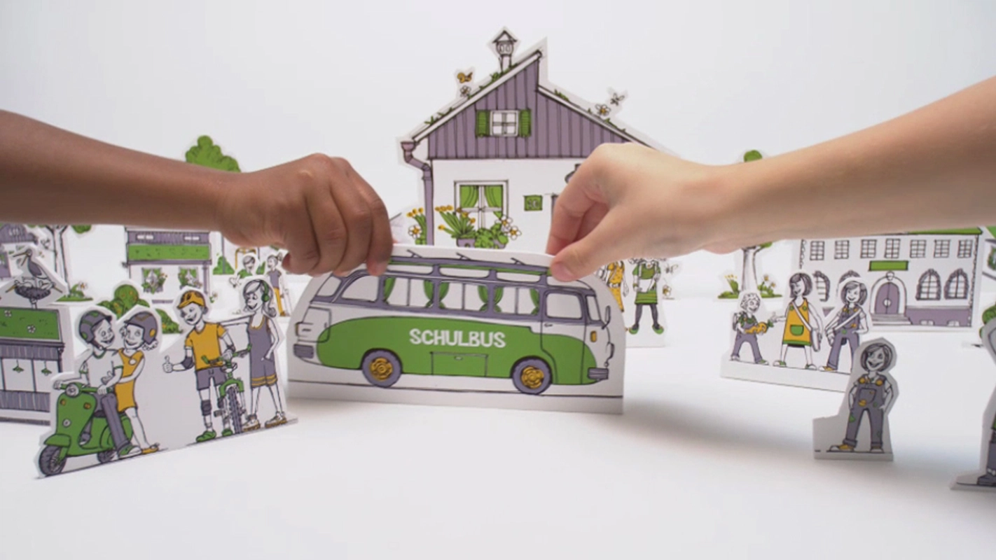 Erklärfilm der Filmproduktion Panda Pictures aus München und Ulm für SOS Kinderdorf. Zwei Hände platzieren einen Schulbus-Modell in einer Szene mit stilisierten Häusern und Figuren.