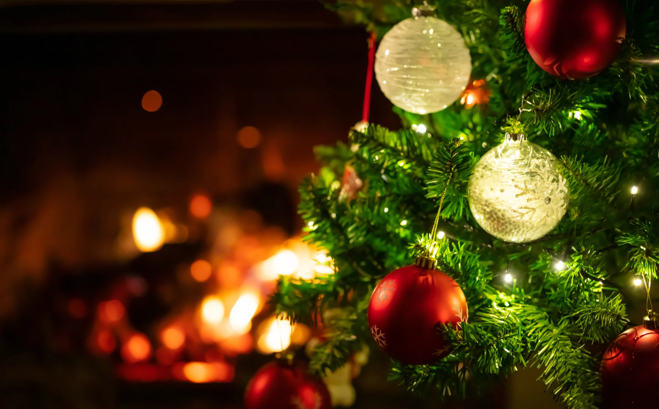 Nahaufnahme von roten und weißen Weihnachtskugeln an immergrünen Zweigen mit dem warmen Glühen eines Kamins, das saisonale Weihnachtswerbung-Themen durch Panda Pictures' Filmproduktion und Videoproduktion in München und Ulm hervorruft.