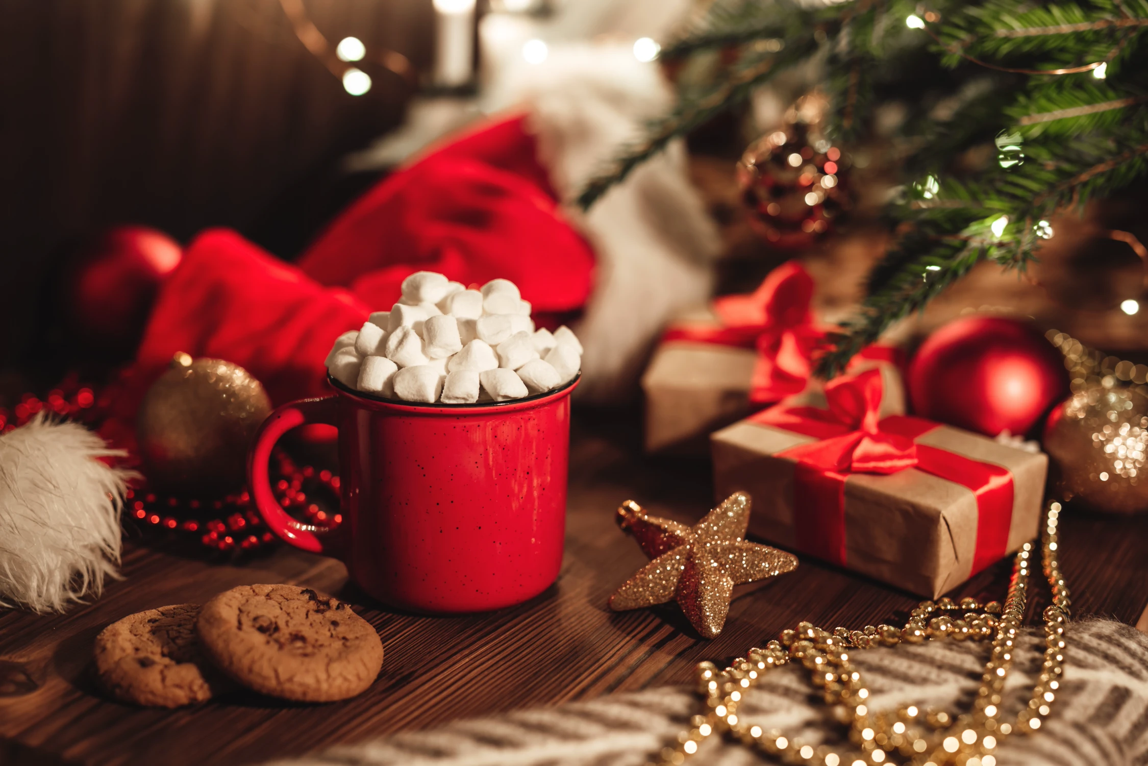 Roter Becher mit Marshmallows, umgeben von Keksen, Geschenken und goldenen Weihnachtsornamenten auf einem Holztisch, ideal für warme Weihnachtswerbung-Bilder von Panda Pictures' Filmproduktion und Videoproduktion in München und Ulm.