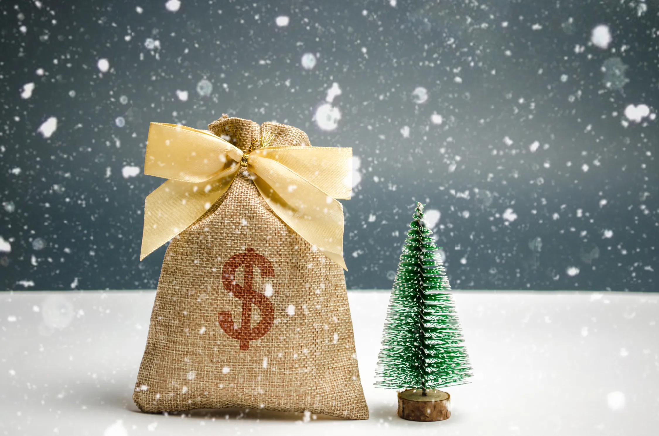 Jutesack mit Dollarzeichen und gelber Schleife neben einem Mini-Weihnachtsbaum in einer verschneiten Umgebung, das Budgetierung von Weihnachtswerbung von Panda Pictures in Filmproduktion und Videoproduktion in München und Ulm symbolisiert.