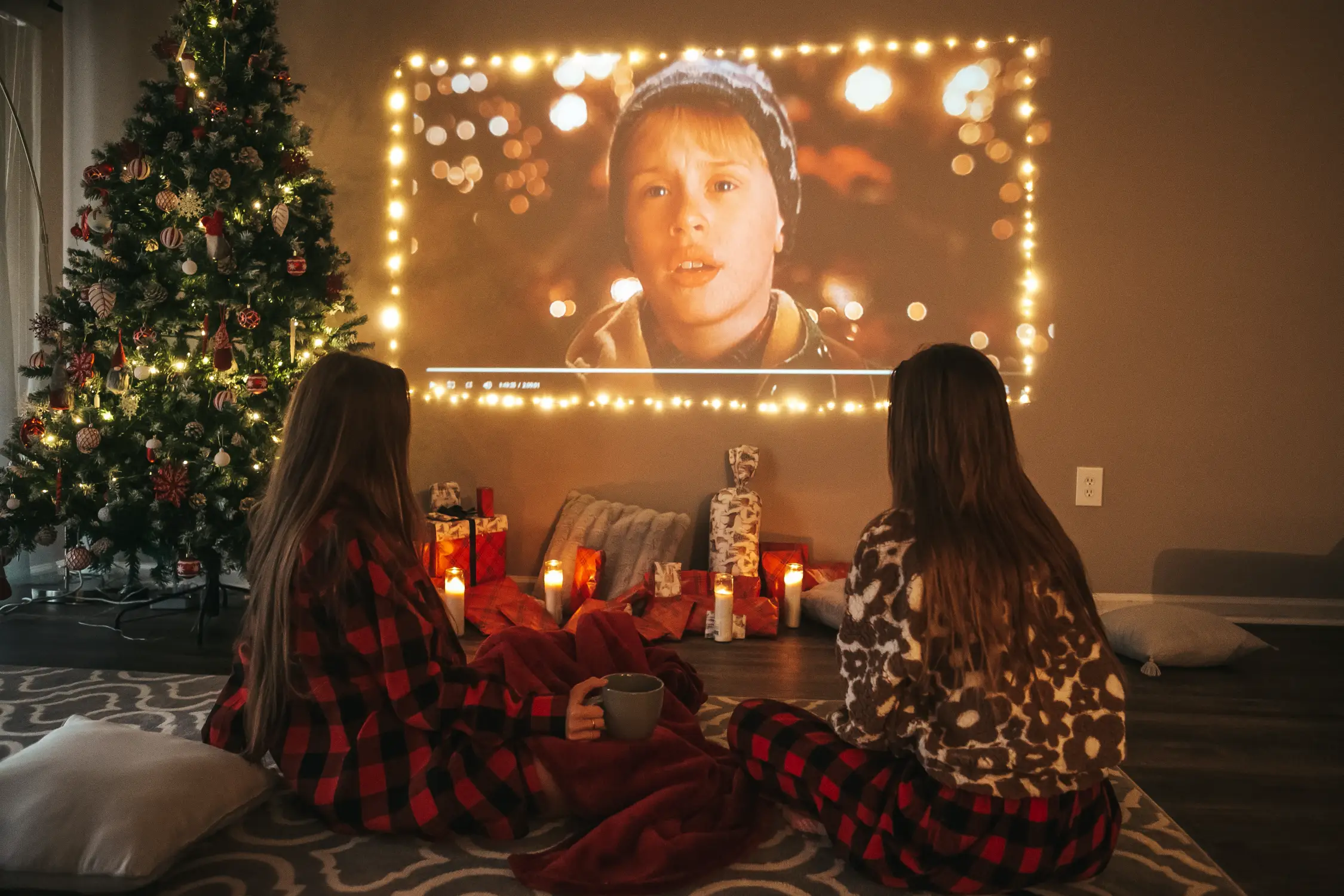 Zwei Personen in Karo-Pyjamas, die vor einem Weihnachtsbaum einen Film auf einer Leinwand betrachten, umgeben von Kerzen und Geschenken, geeignet für Weihnachtswerbung-Atmosphären von Panda Pictures in Filmproduktion und Videoproduktion in München und Ulm.