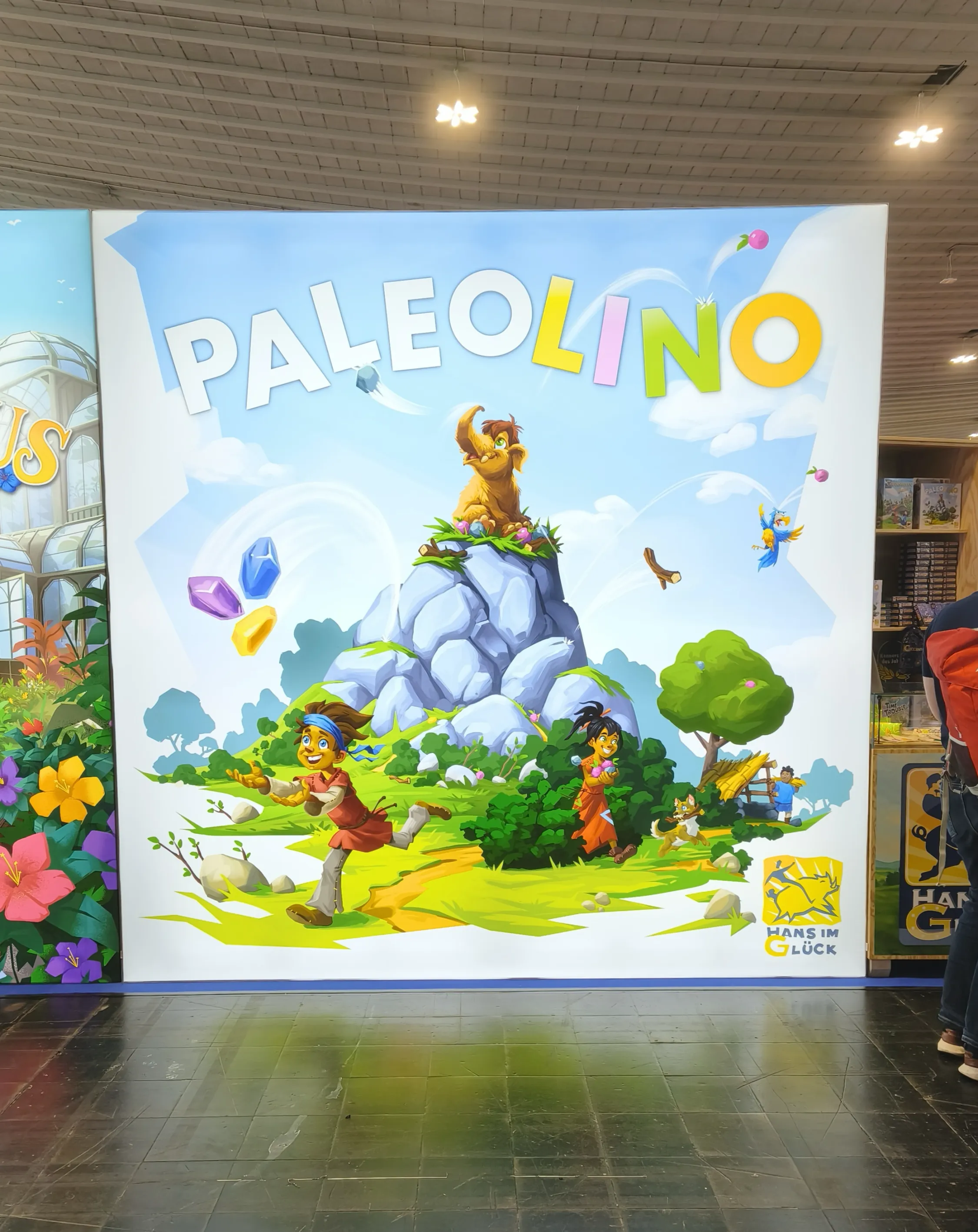 Werbestand für das Brettspiel 'Paleolino' von 'Hans im Glück' auf der SPIEL DOCH! 2026 in Dortmund. Das Banner zeigt ein Mammutbaby auf einem Felsen, Kinder und schwebende Kristalle, aufgenommen für eine Filmproduktion von Panda Pictures.