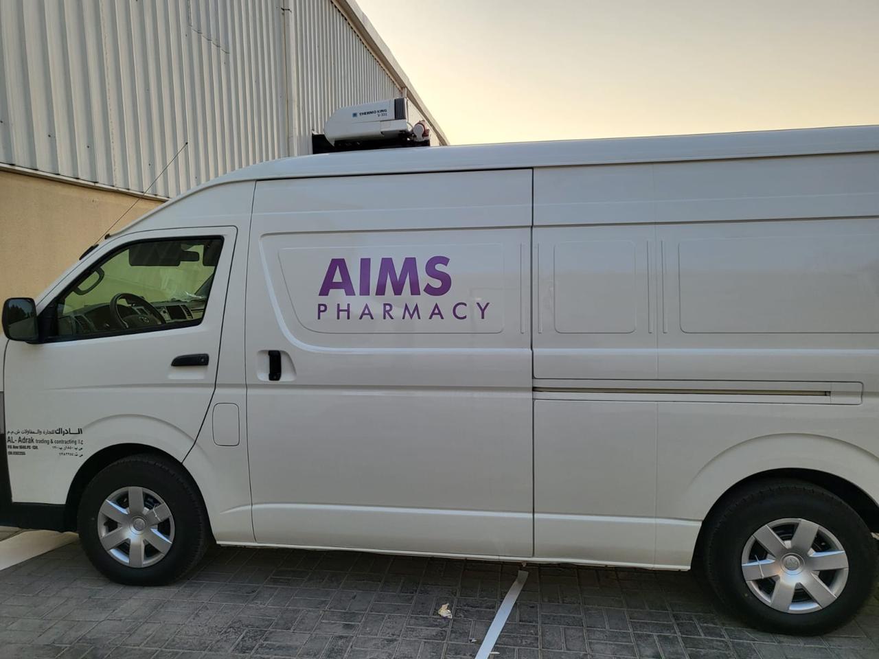White van with "AIMS Pharmacy" logo parked next to a building.