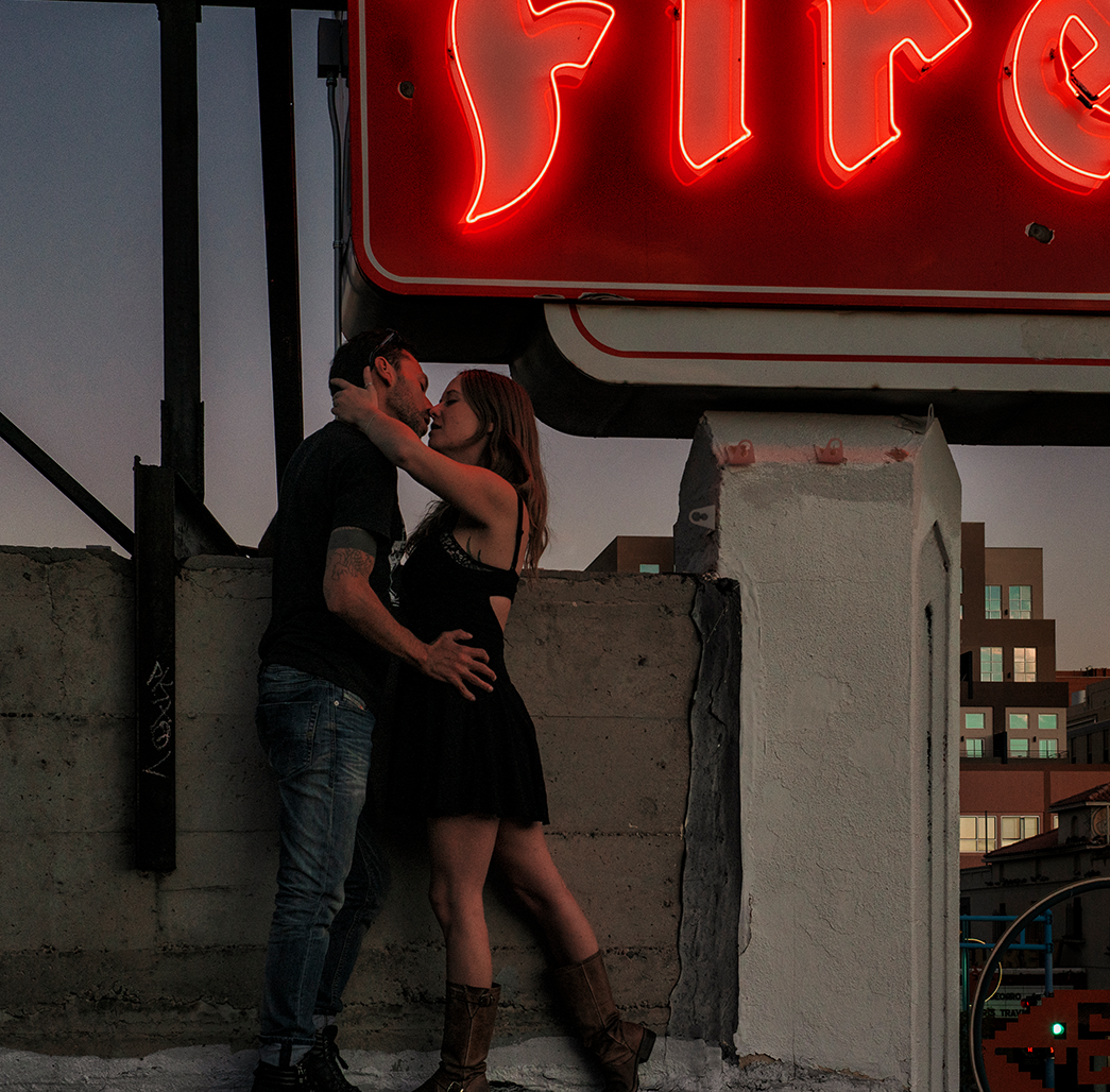 Couple embracing on rooftop at dusk under neon Firestone sign with Albuquerque city skyline in background.