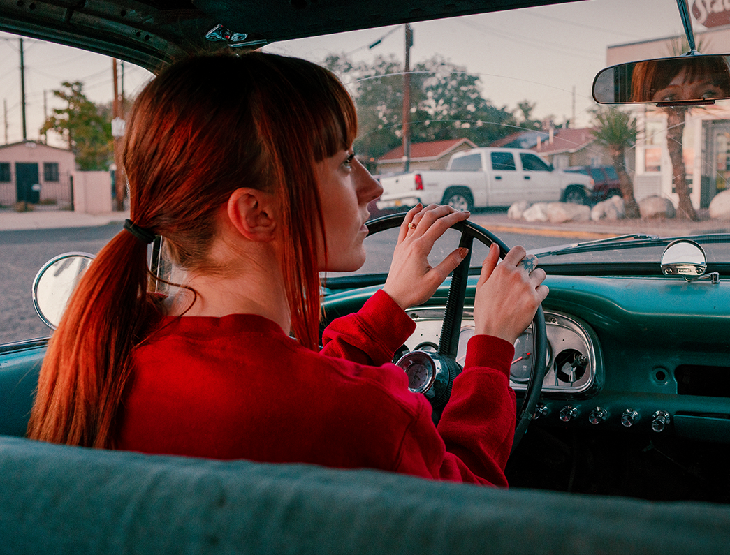 Women wearing red sweater has hands on wheel of a 50s model car, driving in parking lot towards liquor store.