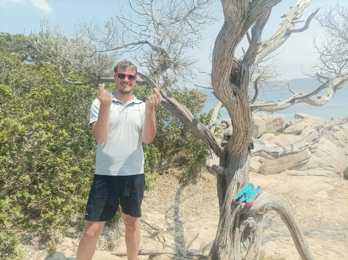 Portrait de l’auteur du blog sur une plage, lunettes de soleil roses, près d’un arbre aux branches torsadées