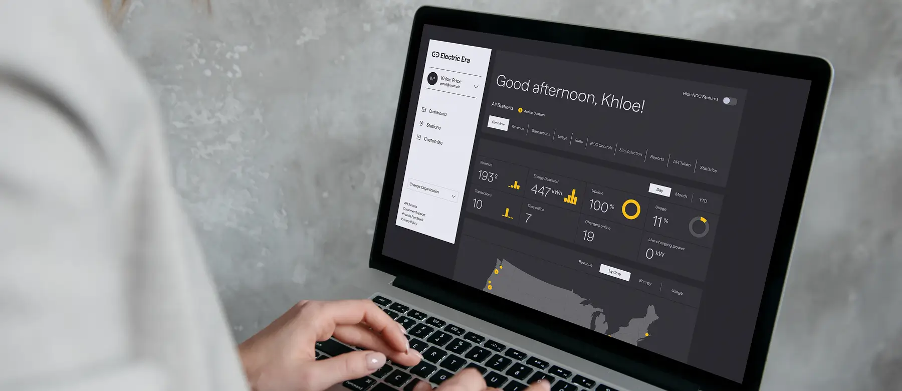 A laptop screen displaying the Electric Era dashboard with detailed energy usage metrics, station information, and system statistics. The interface shows a greeting, 'Good afternoon, Khloe!' along with various charts and data, including energy delivered, u