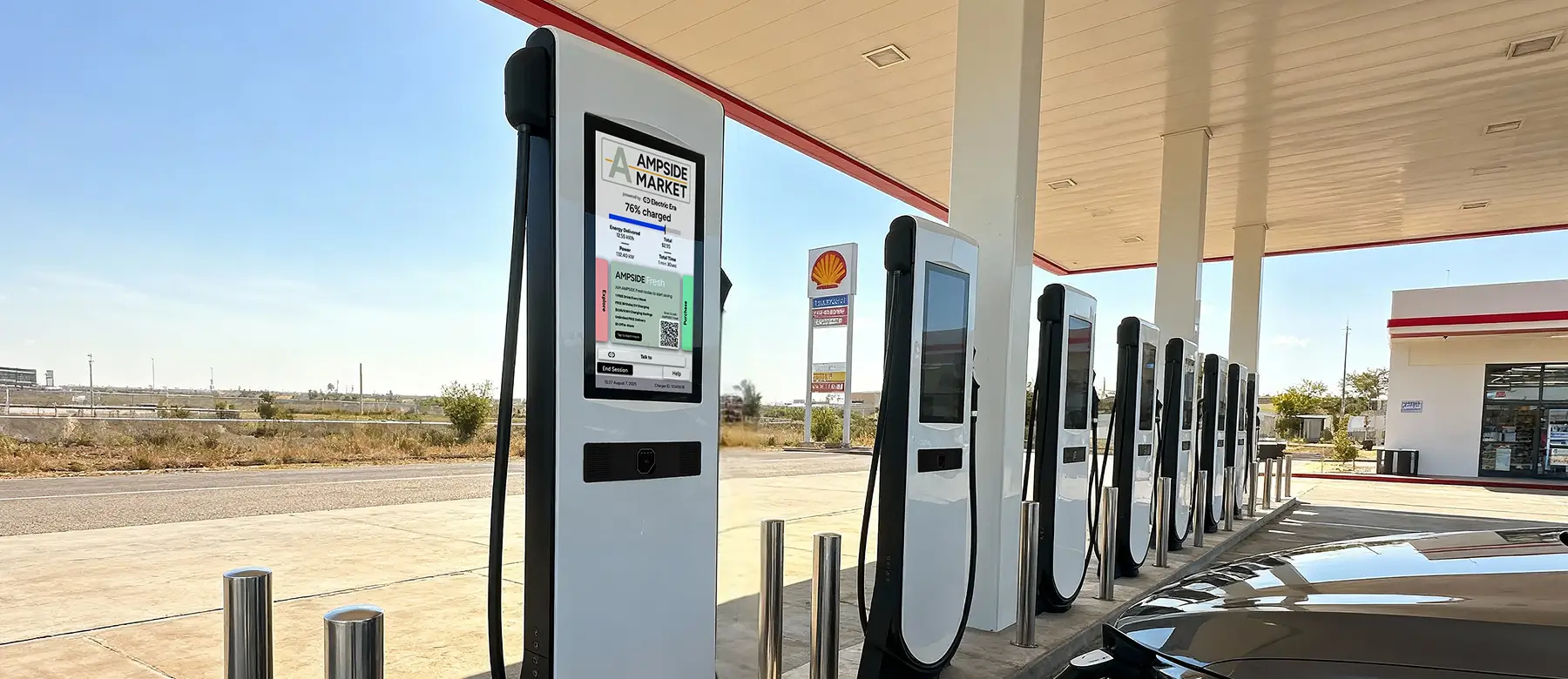 Electric vehicle charging station with a 76% charge displayed on the screen, located at a service station with multiple other charging stations visible in the background. The Shell logo is visible on the building in the background
