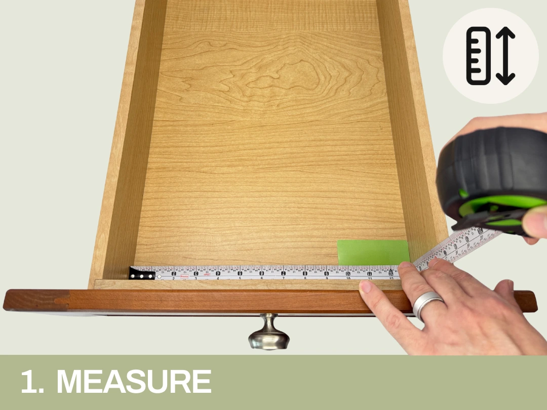 Person using a tape measure to record the interior width of an empty wooden drawer, with a label that reads "1. MEASURE" at the bottom and an icon of a ruler with vertical arrows in the top right corner. 