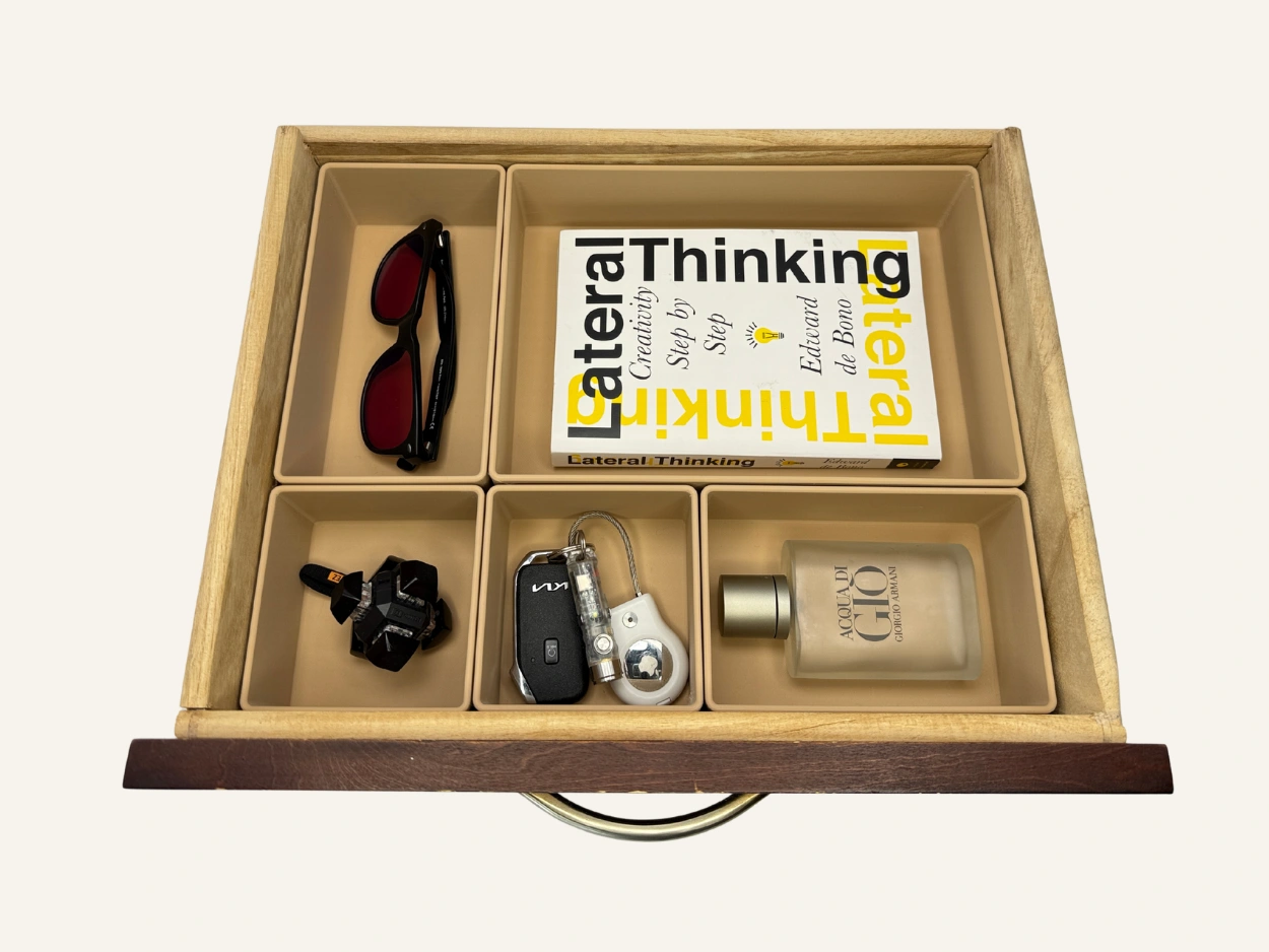 Open drawer with six beige compartments holding organized items including a pair of sunglasses, a book titled Lateral Thinking, a small flashlight, a keychain with car keys and a flashlight, and a bottle of cologne.