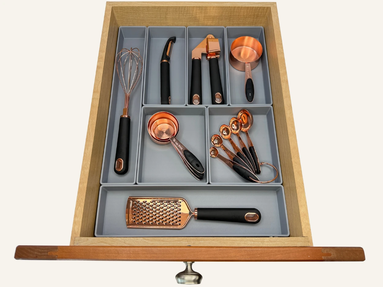 Open drawer with gray custom compartments holding organized copper and black kitchen tools, including a whisk, vegetable peeler, garlic press, measuring cups and spoons, a grater, and a ladle.