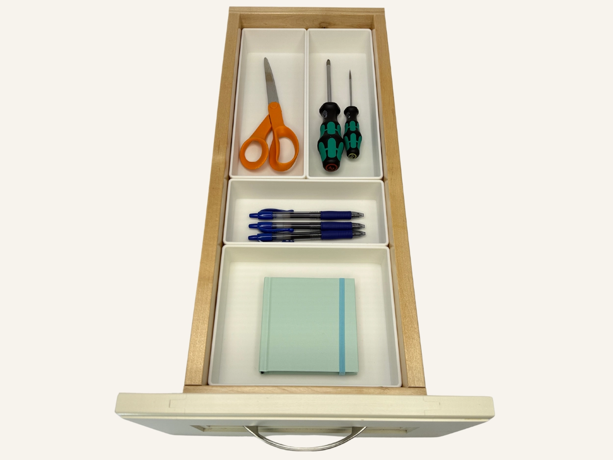 Open drawer with white custom compartments containing orange scissors, two green-handled screwdrivers, four blue pens, and a closed light blue notebook.