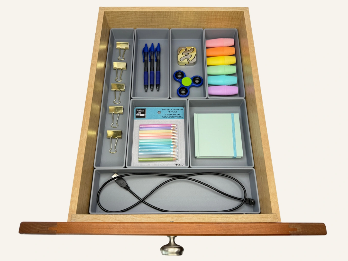 Open drawer with gray custom compartments holding binder clips, blue pens, puzzle piece, colorful highlighters, fidget spinner, pastel colored pencils, sticky notes, and a black USB cable.