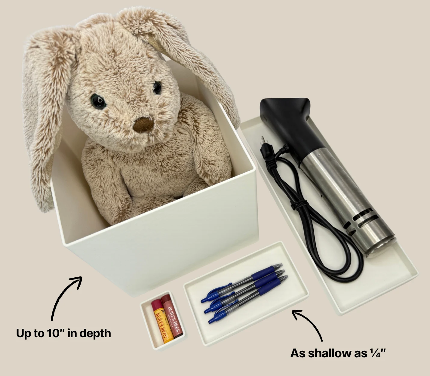 Custom white storage containers of varying depths displayed on a beige background. One deep container holds a plush bunny, while shallow containers hold items like pens, lip balm, and a sous vide machine. Text labels indicate depths ranging from ¼ inch to 10 inches.