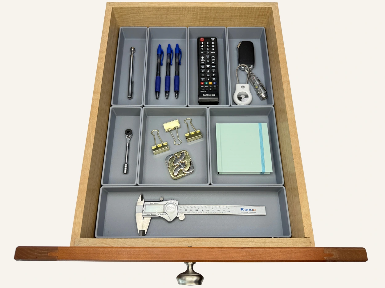 Open drawer with gray custom organizers neatly holding various household and office items, including pens, a remote control, car keys, sticky notes, a wrench, gold binder clips, a puzzle piece, and a digital caliper.