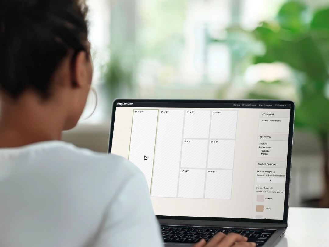 Woman using a laptop with the AnyDrawer design tool open on the screen, showing a custom drawer layout with divider dimensions and customization options.