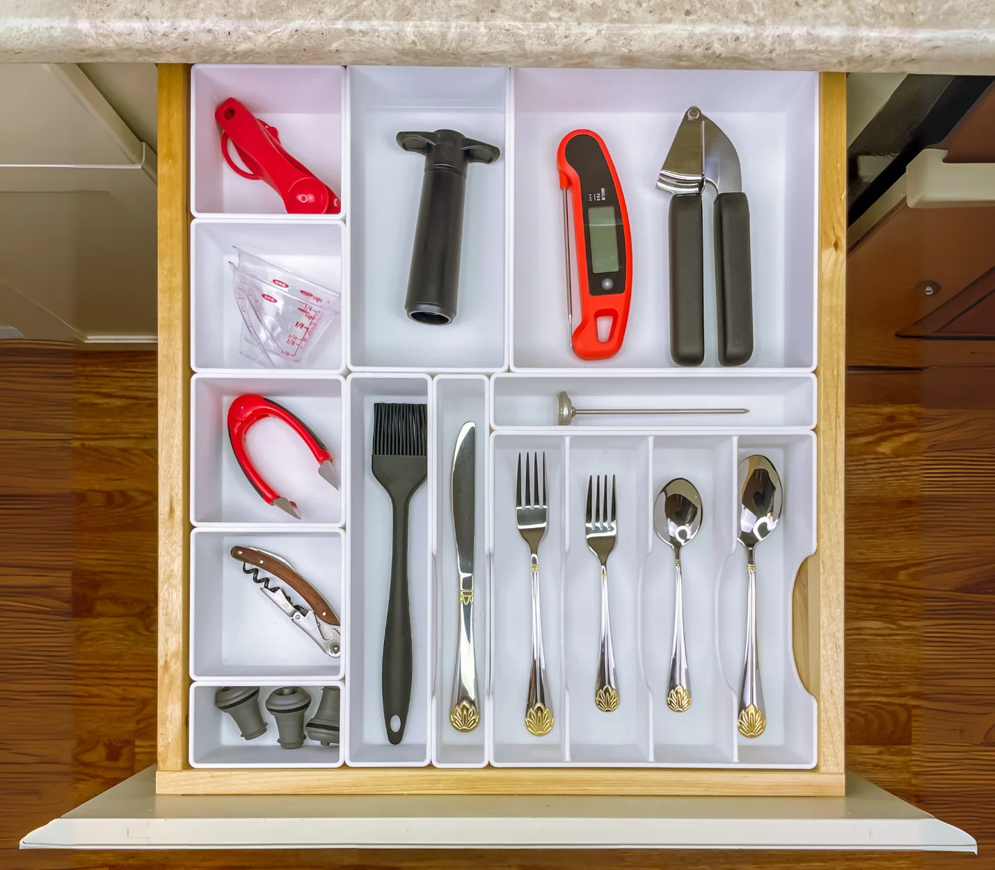 Top-down view of a kitchen drawer with white custom trays holding utensils, a garlic press, thermometer, measuring tools, and various kitchen gadgets.