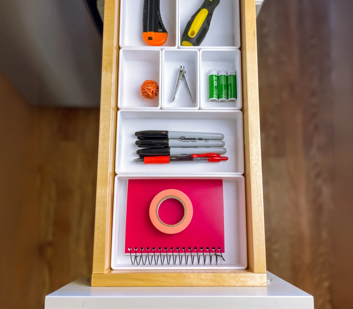 Top-down view of a junk drawer with white custom trays organizing pens, Sharpies, a rubber band ball, tape, a notebook, batteries, and utility tools.