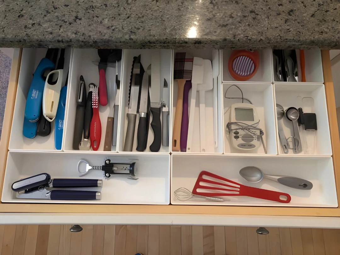 Top-down view of a kitchen drawer organized with white custom compartments holding various utensils including lighters, peelers, knives, spatulas, a digital thermometer, measuring spoons, and a corkscrew.