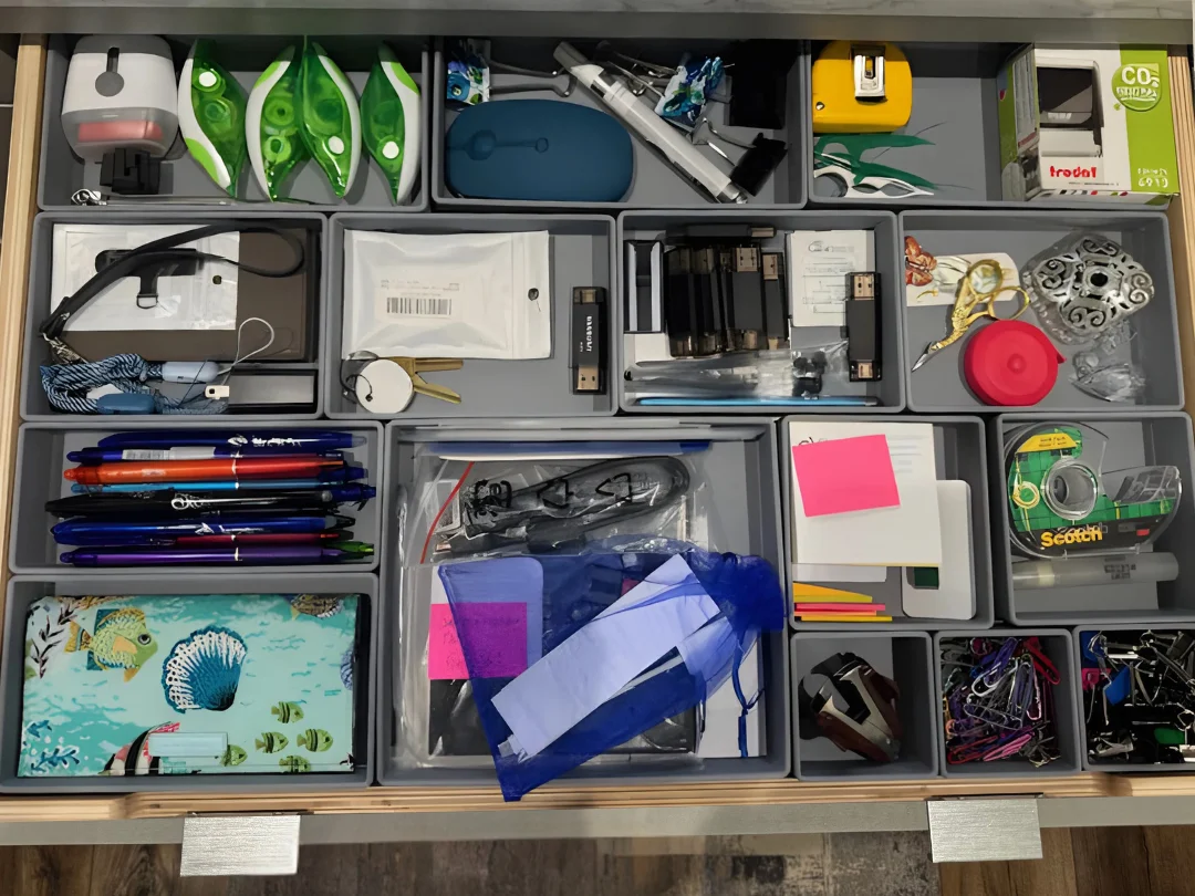 Top-down view of a gray custom-organized drawer containing various office supplies including pens, scissors, USB drives, correction tape, chargers, sticky notes, tape rolls, binder clips, and a decorative fish-patterned item.