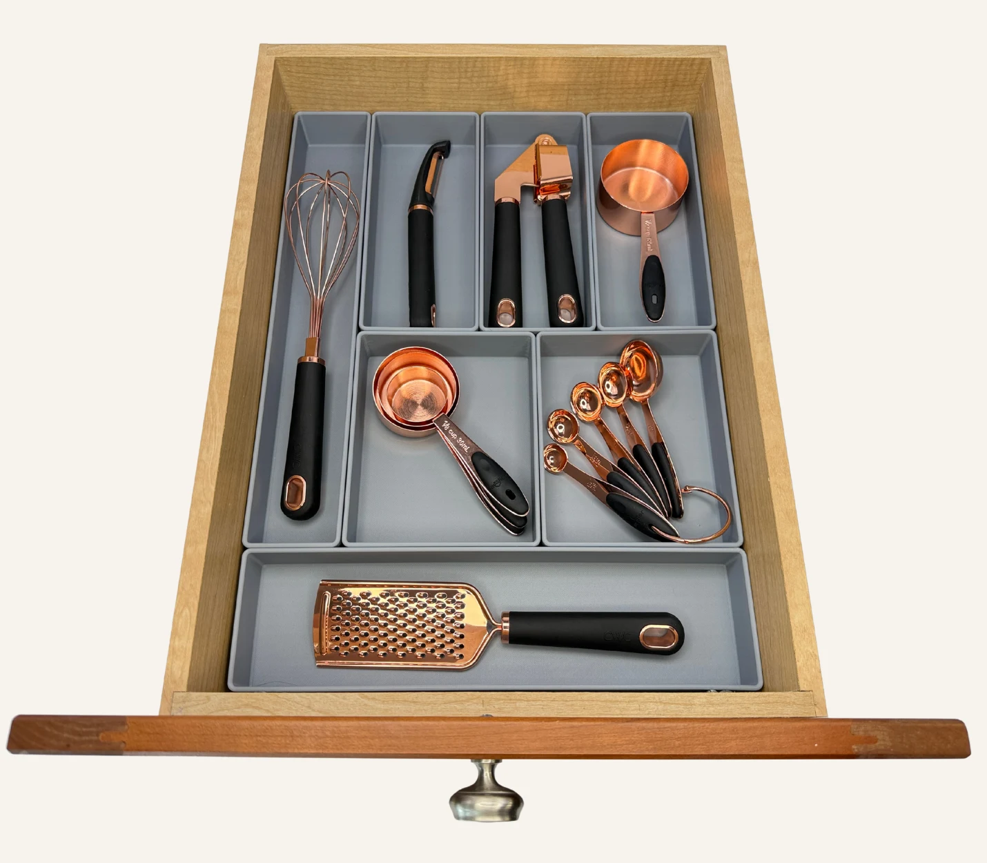 Drawer with gray dividers neatly organizing copper and black kitchen utensils, including a whisk, peeler, garlic press, measuring cup, measuring spoons, grater, and scoops.