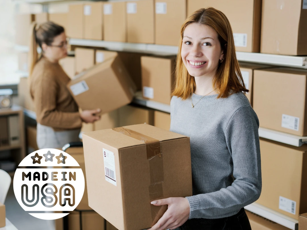 Smiling woman holding a cardboard box in a warehouse filled with stacked boxes, with a “Made in USA” badge overlaid in the lower left corner.
