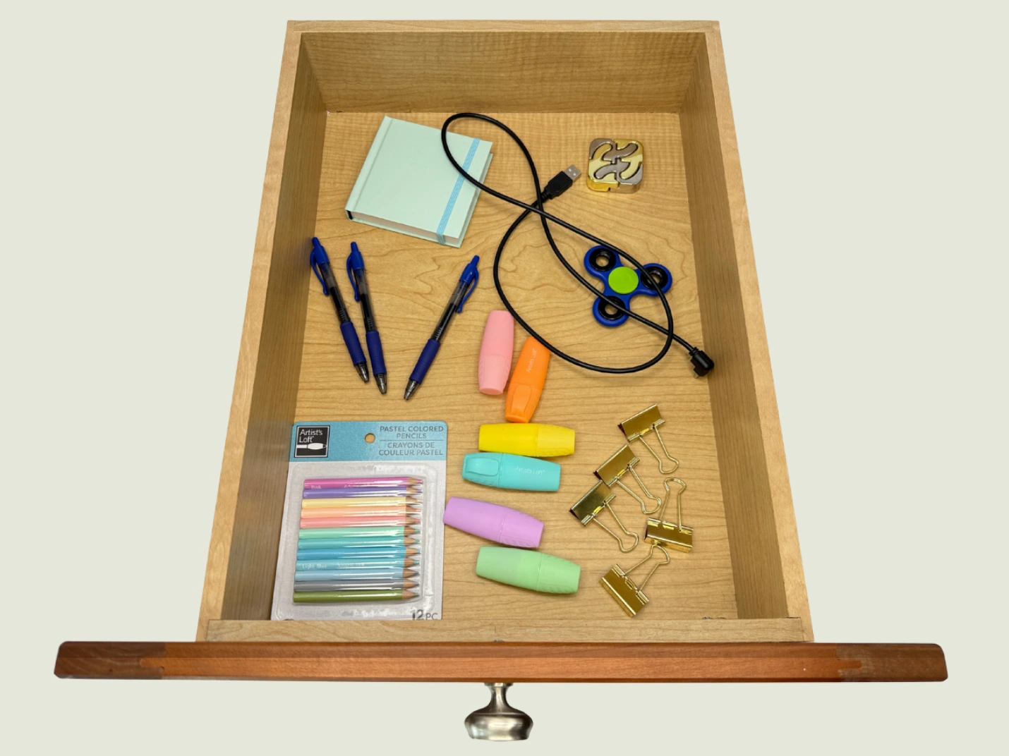 Open drawer with white custom compartments containing orange scissors, two green-handled screwdrivers, four blue pens, and a closed light blue notebook.