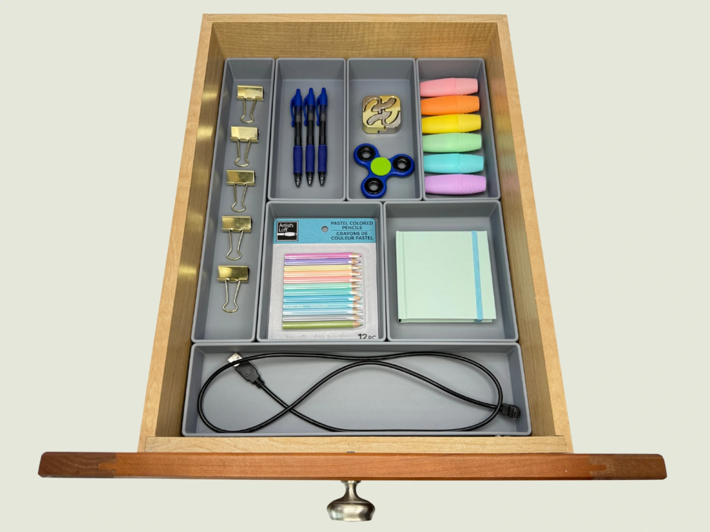 Open drawer with gray custom compartments holding binder clips, blue pens, puzzle piece, colorful highlighters, fidget spinner, pastel colored pencils, sticky notes, and a black USB cable.