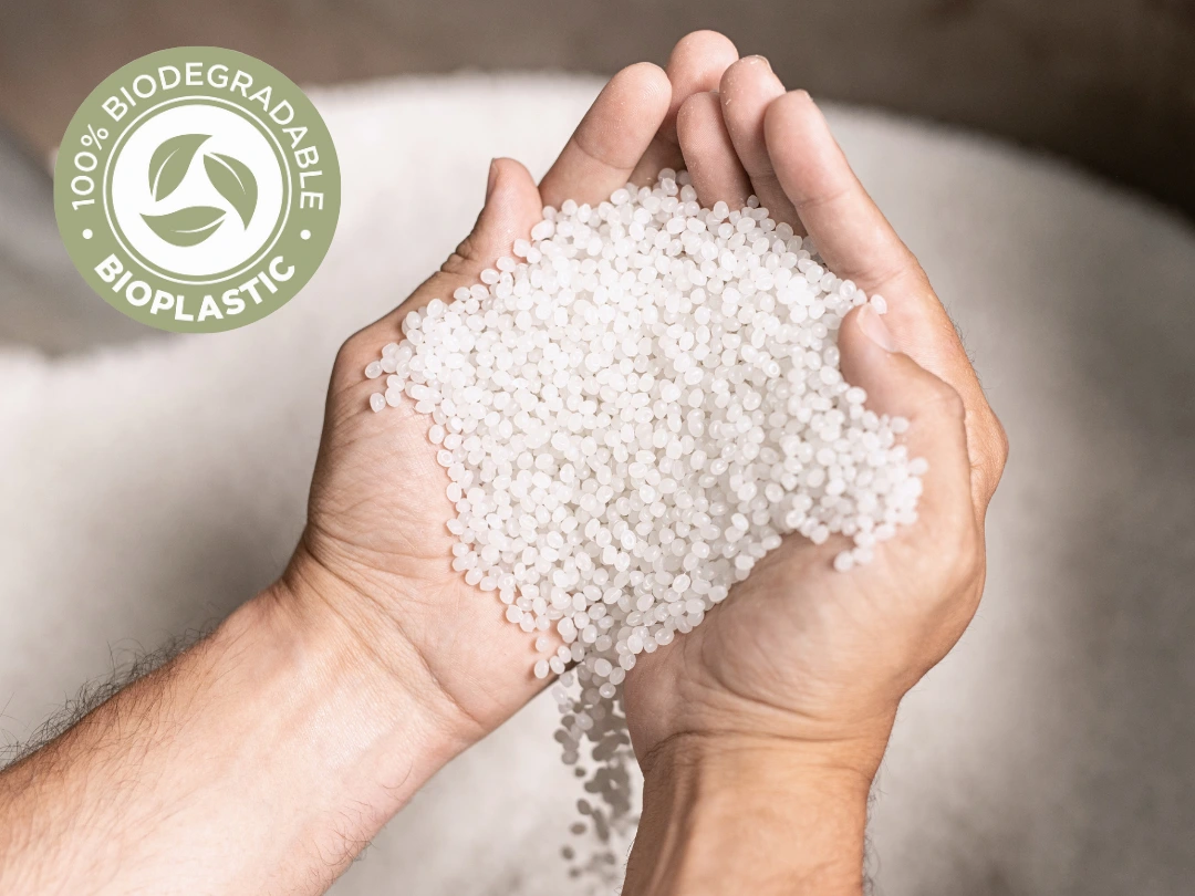 Hands holding a pile of small white bioplastic pellets, with a circular badge in the top left corner that reads “100% Biodegradable Bioplastic.”