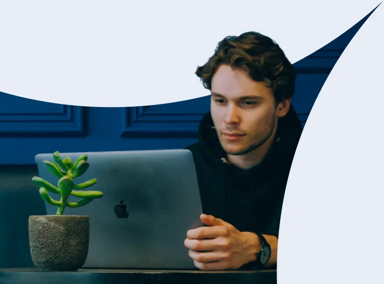 A man is sitting in front of a laptop with a plant in a pot on the table.