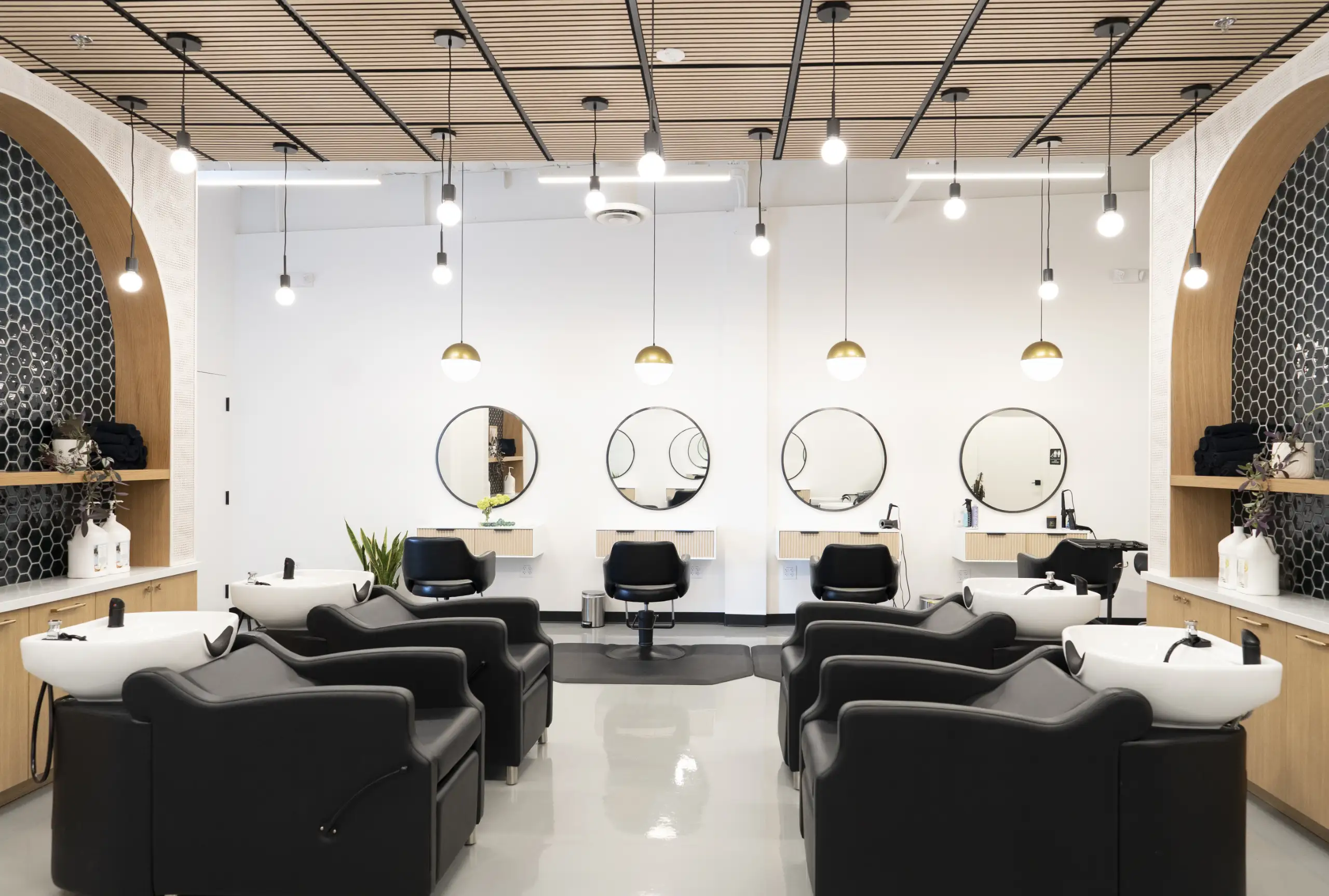 Modern hair salon interior with black wash chairs and sinks on both sides and styling chairs with round mirrors on a white wall.