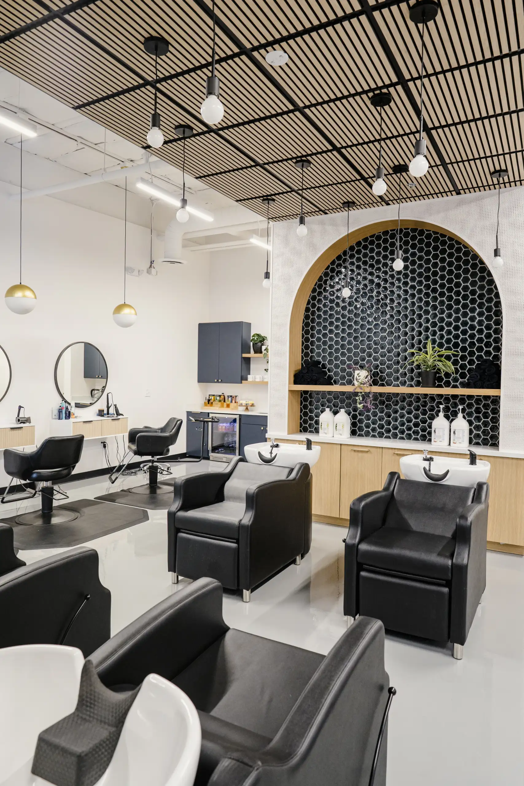 Modern hair salon interior with black styling chairs, white wash basins, wooden ceiling panels, and a black hexagonal tiled wall with an arched wooden frame.