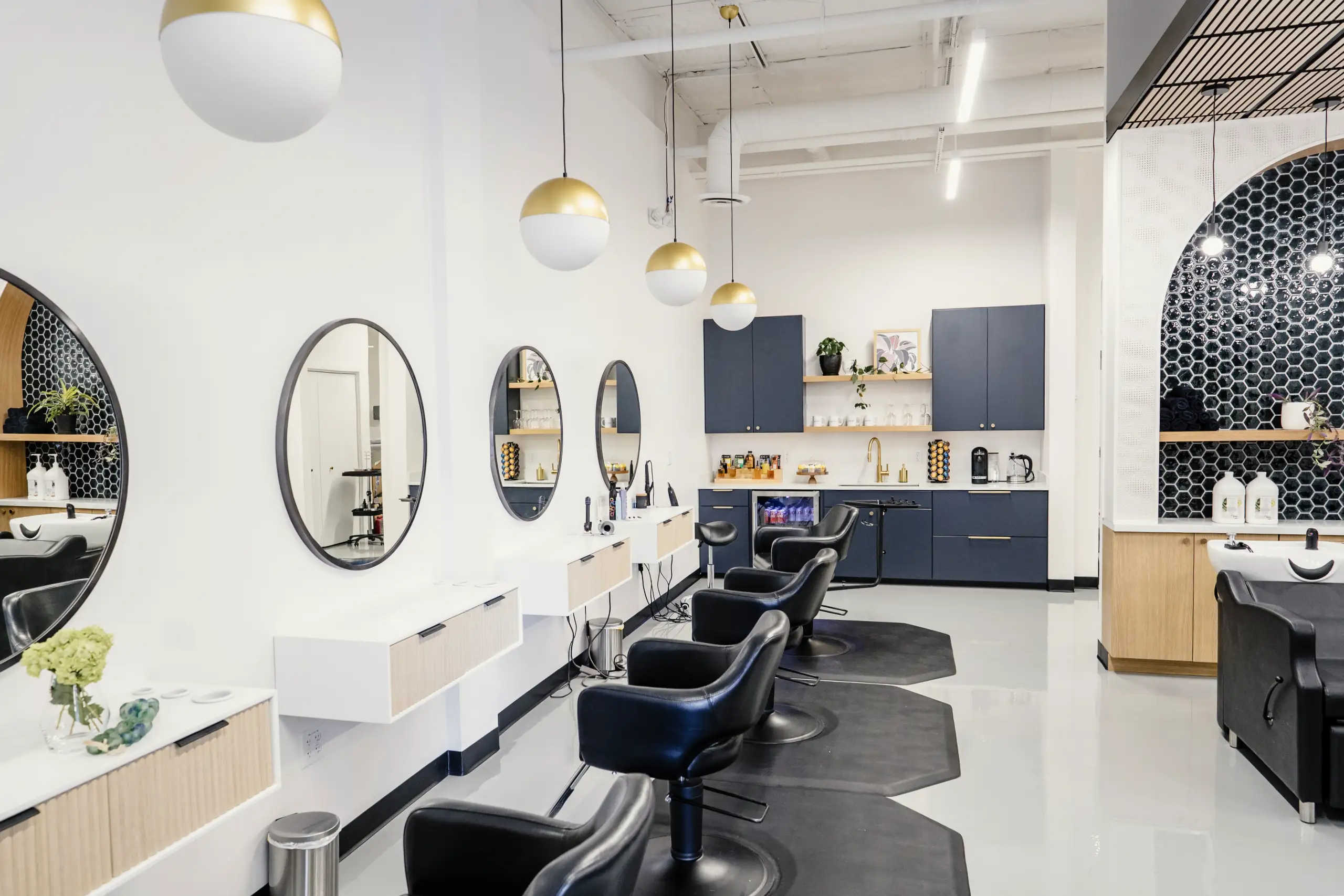 Modern hair salon interior with four black styling chairs facing oval mirrors, white walls, gold and white hanging lights, and a back area with dark blue cabinets and hair care products.