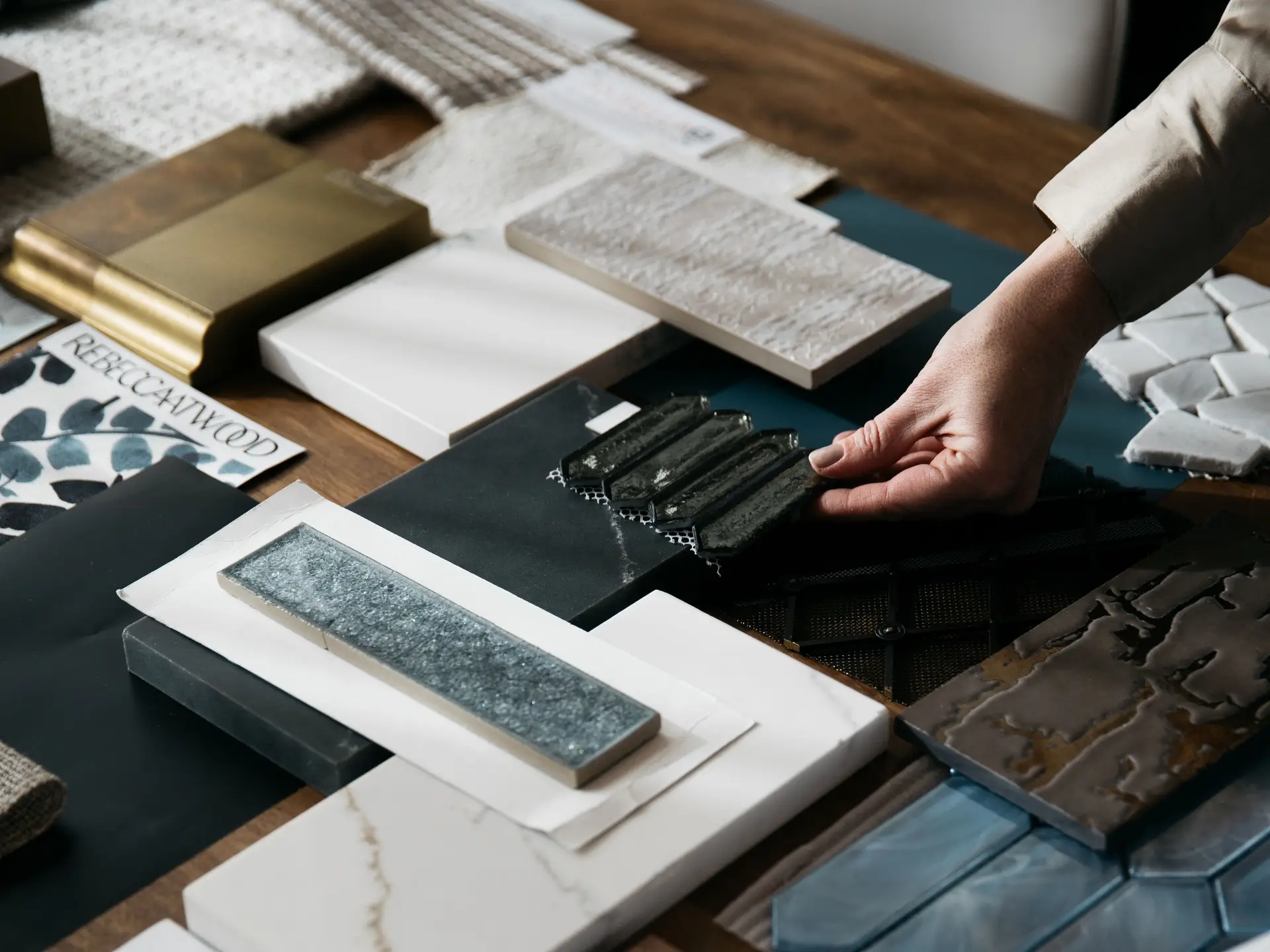 Person arranging various tile and fabric samples on a wooden table for interior design.