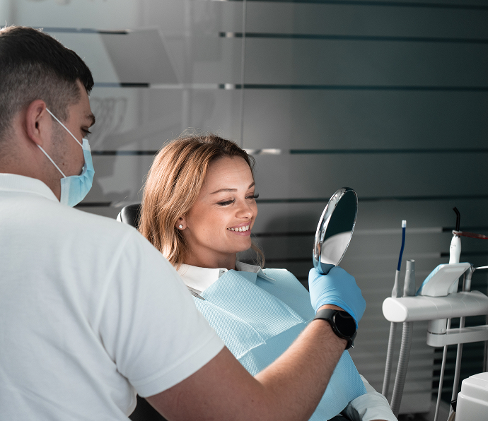 A masked dentist holding a mirror to a patient examining her smile after a dental procedure. 