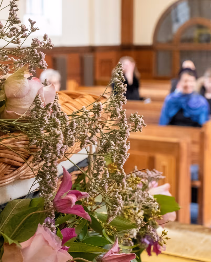 A close up of a spray of flowers on a coffin with mourners sat in pews in the background. Photo by Good Funeral Guide on Unsplash. 