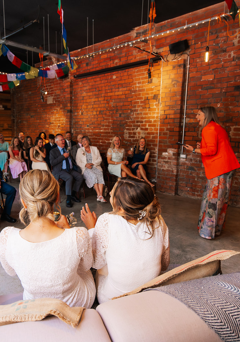 Wedding celebrant Kate Rostance leading Shell and Jenna’s industrial-style ceremony in front of family and friends, with laughter and warmth filling the brick-walled venue.